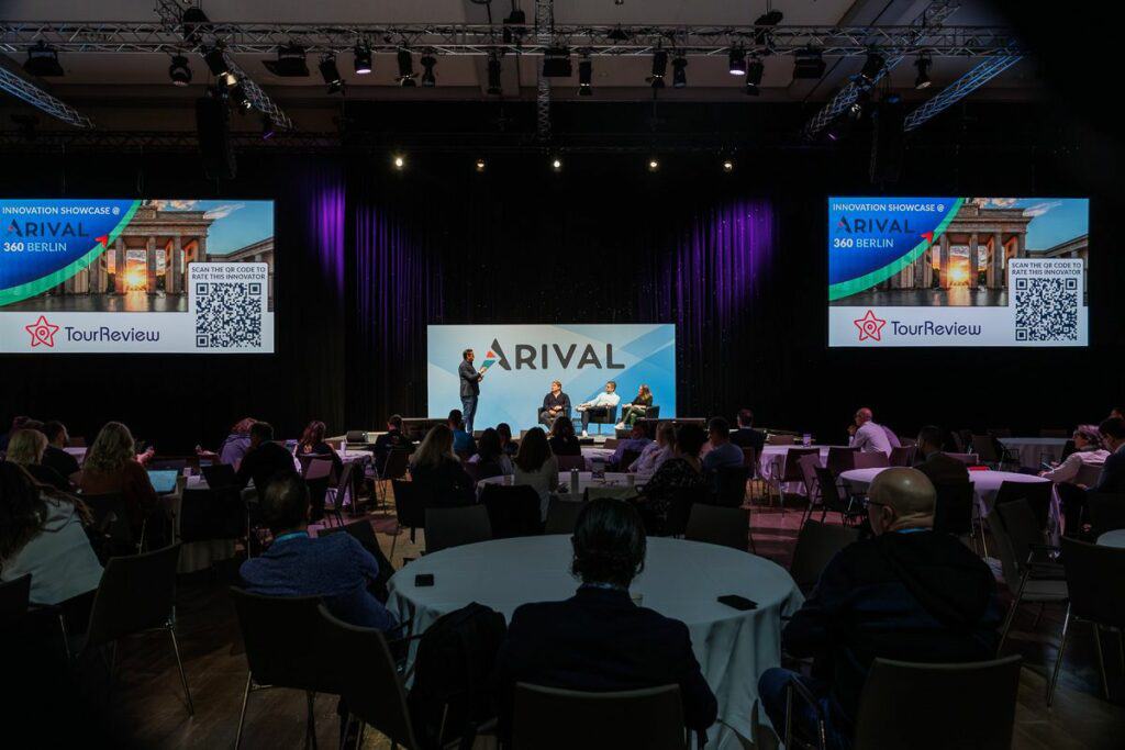 Conference hall with audience watching a presenter on stage at the Arival event. Large screens display TourReview logos and QR codes.