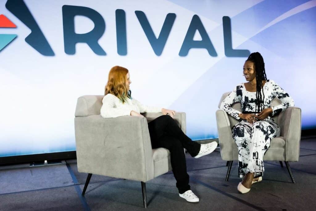 Two women seated on a stage having a conversation, with "ARIVAL" logo displayed prominently in the background.