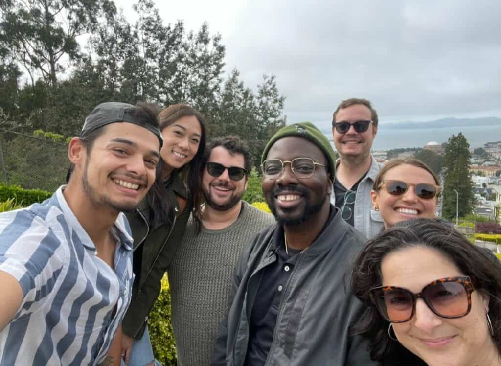 Group selfie of seven diverse people smiling outdoors with trees and overcast sky behind them.