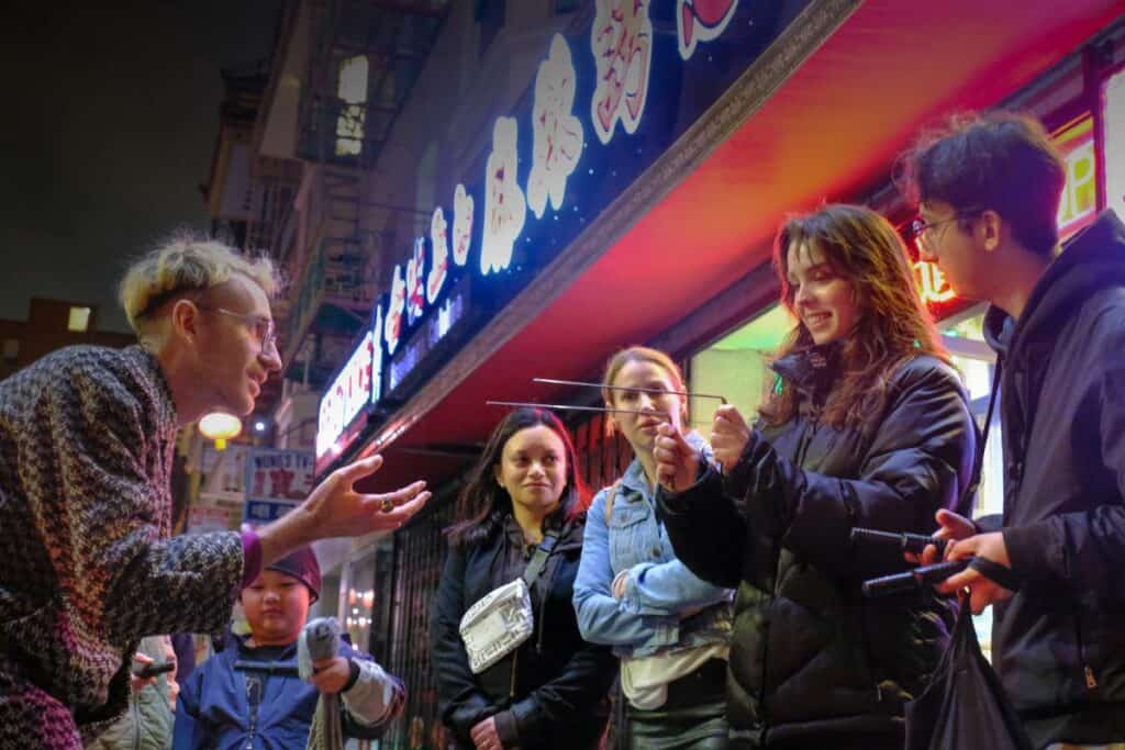 Tour guide gesturing while speaking to a diverse group of people at night, in front of a colorful storefront.