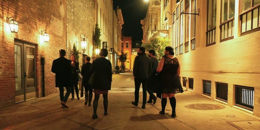 Nighttime street scene with people walking along a well-lit alley between brick buildings.