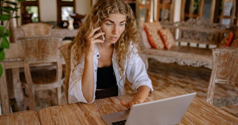 Young woman with curly blonde hair talking on phone while typing on laptop at wooden table in bright, modern interior space.