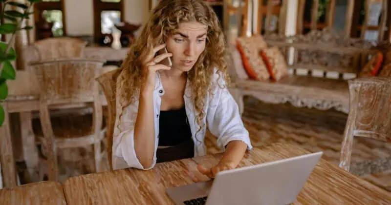 Young woman with curly blonde hair talking on phone while typing on laptop at wooden table in bright, modern interior space.