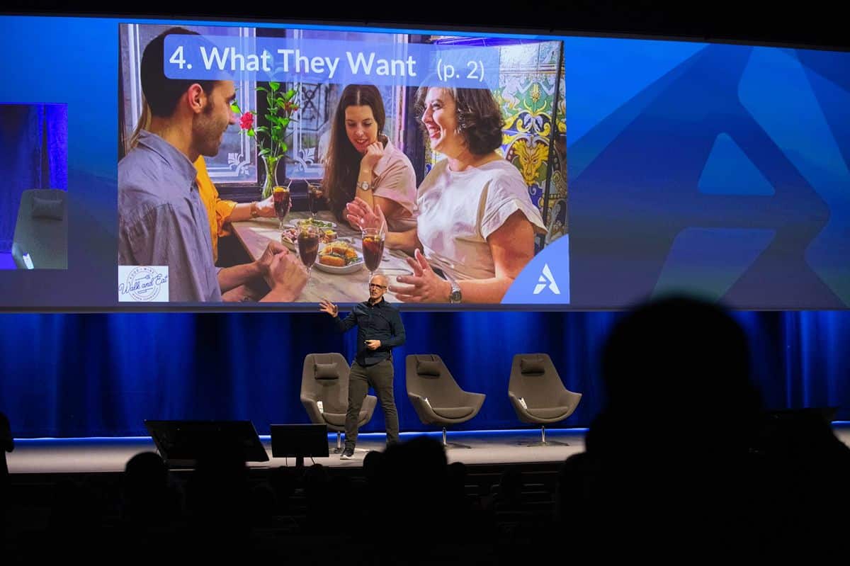 A speaker gestures on stage at an Arival event beneath a large screen showing a slide titled "4. What They Want," with a photo of travelers laughing over drinks and tapas on a Walk and Eat food tour.