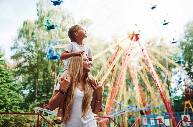 Daughter sits on the shoulders. Cheerful little girl her mother have a good time in the park together near attractions.
